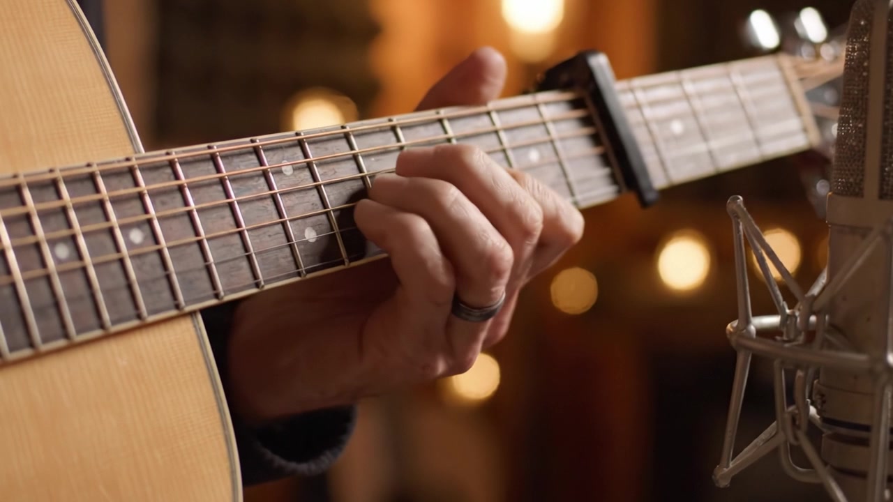 Una vista ravvicinata di una mano che strimpella abilmente accordi su una chitarra acustica su uno sfondo caldo e avvolgente illuminato da luci soffuse.