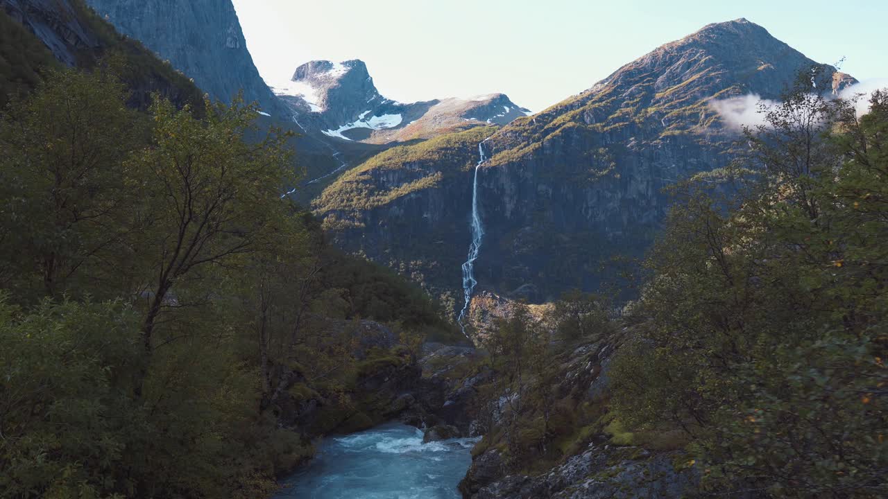 Kamera se pomalu pohybuje panoramaticky otevřeným lesem s krásnou řekou a vodopádem stékajícím z hory v pozadí