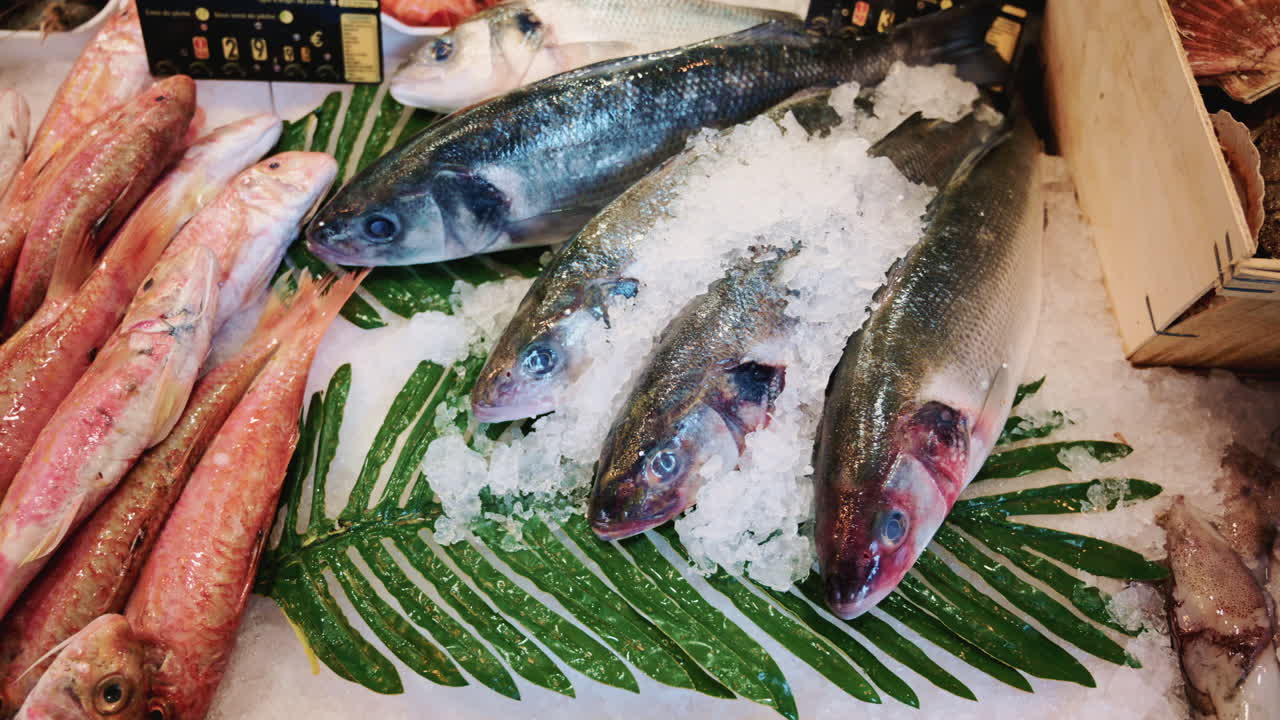 Assortment of whole fish laid on ice with green leaves at a market stall, showing variety and abundance of fresh seafood