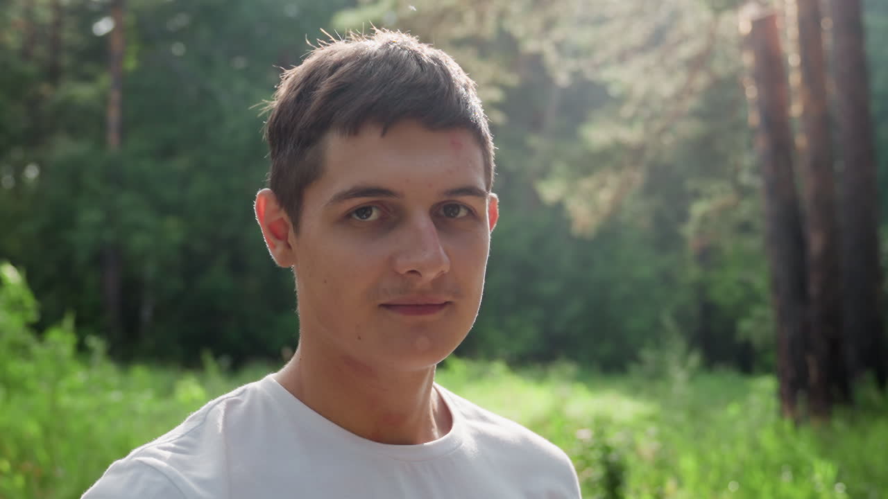 Portrait of handsome young man in white shirt standing in soft sunlight with calm expression, surrounded by green nature and forest background, peaceful atmosphere capturing serenity