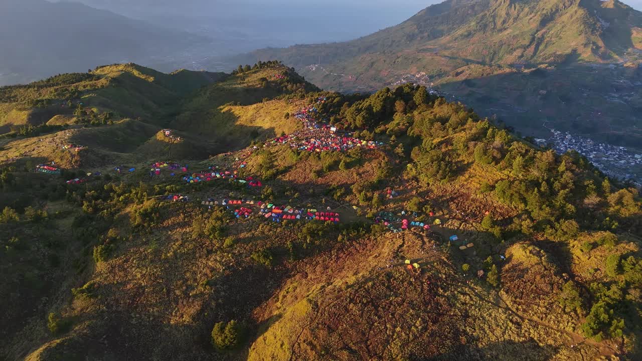 Drone footage showcasing a massive camping site spread across rolling mountain hills, with vivid tents glowing in the warm morning light. Sunrise camp point of Mount Prau, Indonesia
