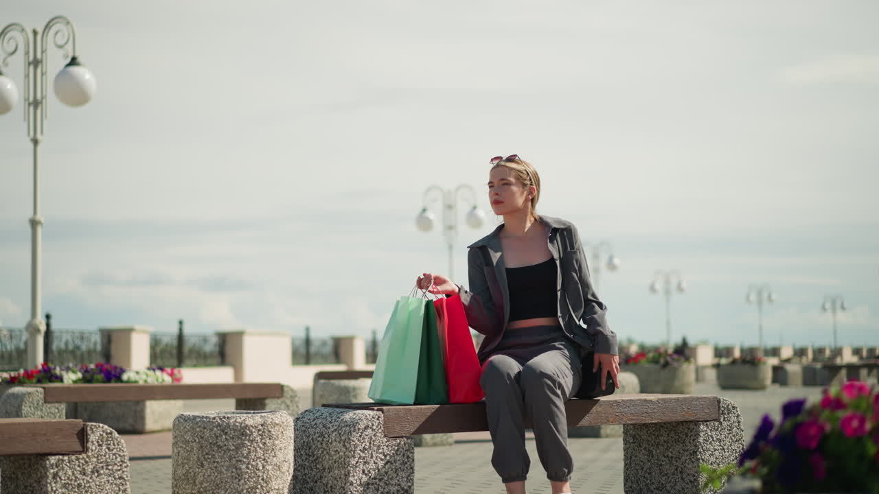 세 개의 쇼핑 가방을 들고 있는 여인이 야외 벤치를 향해 걸어가며, 벤치에 가방을 조심스럽게 놓고, 앉아서, 검은색 드백을 조정하고, 다리를 가로지르며, 다리를 단합니다.