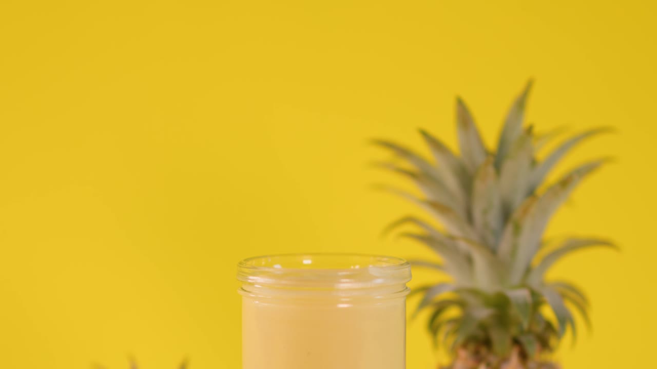 Hand places pineapple slice on creamy smoothie, vibrant yellow backdrop, tropical fruit, bright lighting