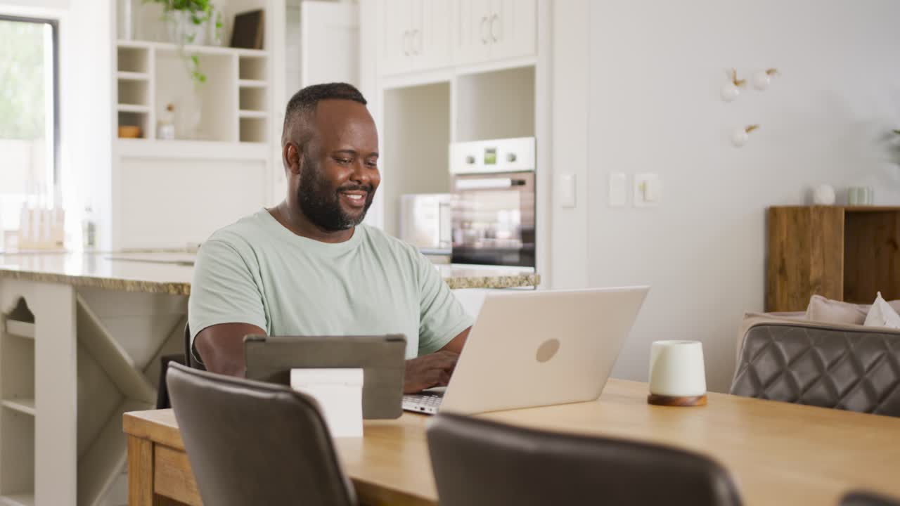 African man typing on laptop checking tablet smiling while reading message at dining table