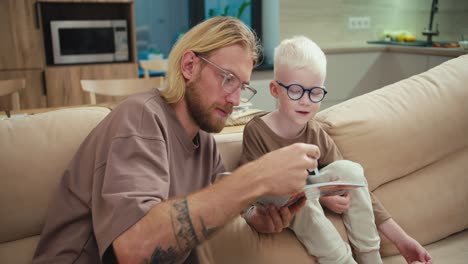 Happy-little-albino-boy-in-blue-glasses-sits-on-the-sofa-and-his-dad,-a-blond-man-with-long-hair-and-a-beard,-explains-to-the-boy-how-to-read-letters-correctly-before-the-first-lessons-at-school-during-home-communication-in-the-kitchen