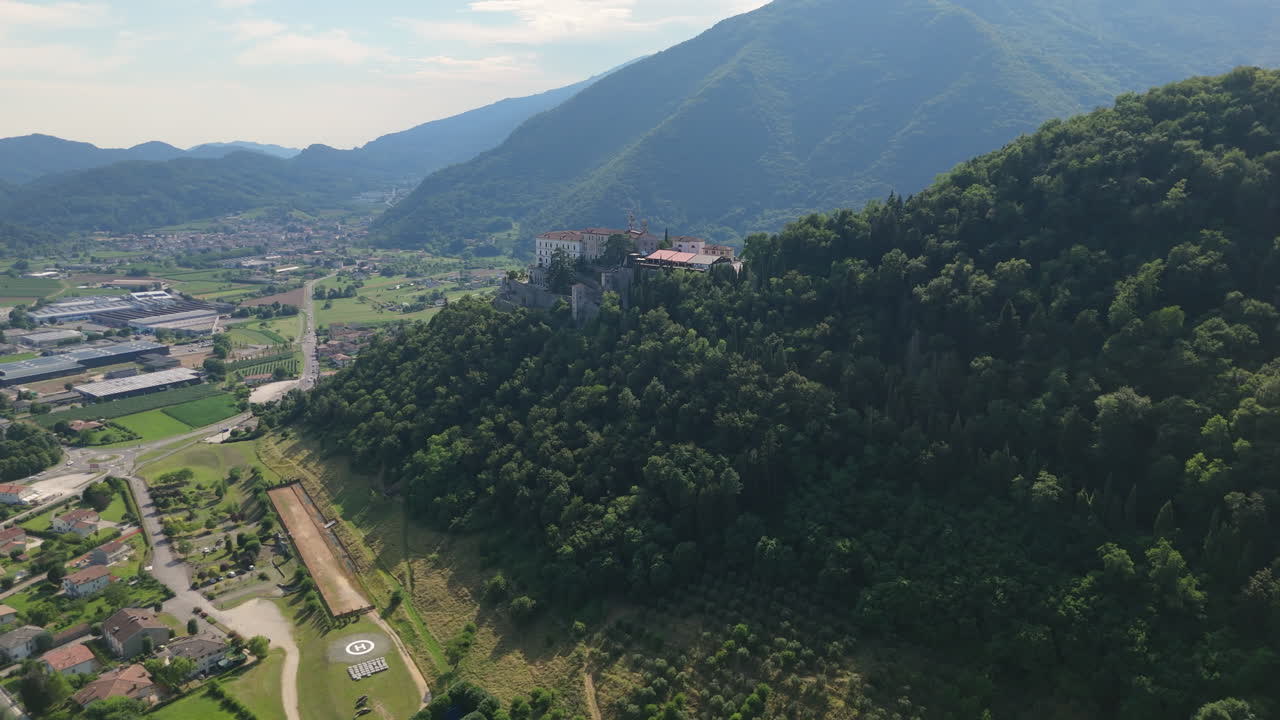Drone view of a historic castle on a forested hill with a wide valley and small towns in the background. Scenic Italian landscape on a sunny day