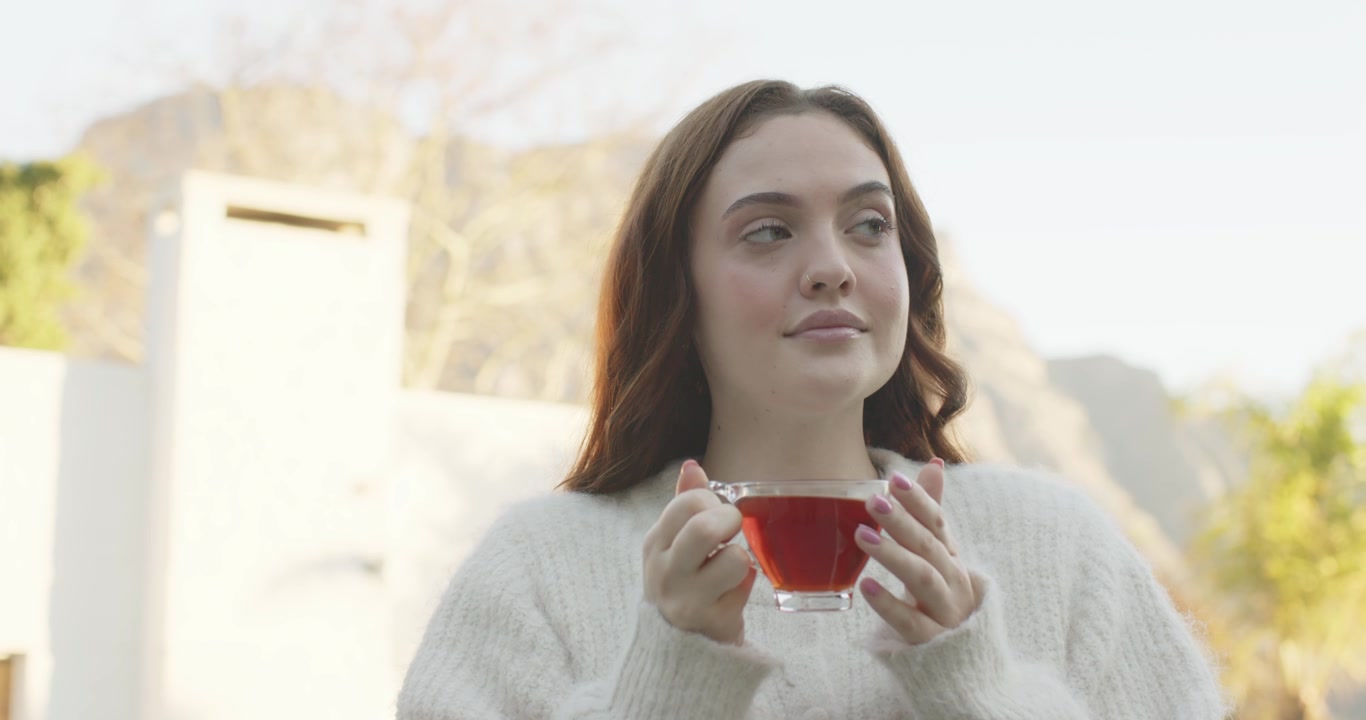 Donna adulta con un maglione morbido su una terrazza, che culla una tazza di tè, inalando l'aroma, assaporando, guardando verso destra.