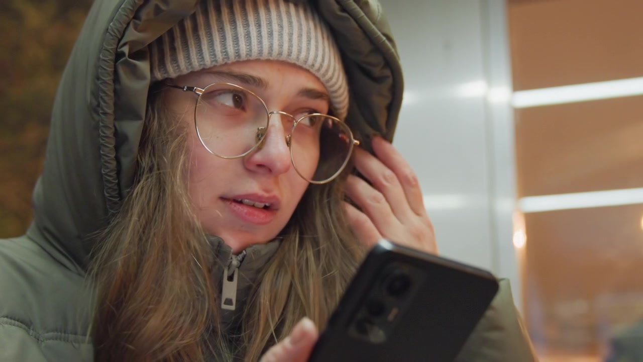 Ung hvid kvinde tjekker telefon med tommelfingeren, der scroller beskeder, skærmglød på brillerne, hættejakke og hue, intimt nærbillede i et transitlæskur, fokuseret tankefuld udtryk