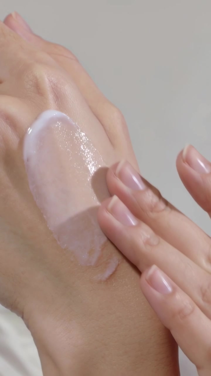 Video vertical: Puntas de los dedos presionando y extendiendo crema nacarada en el dorso de la mano, mostrando un brillo.