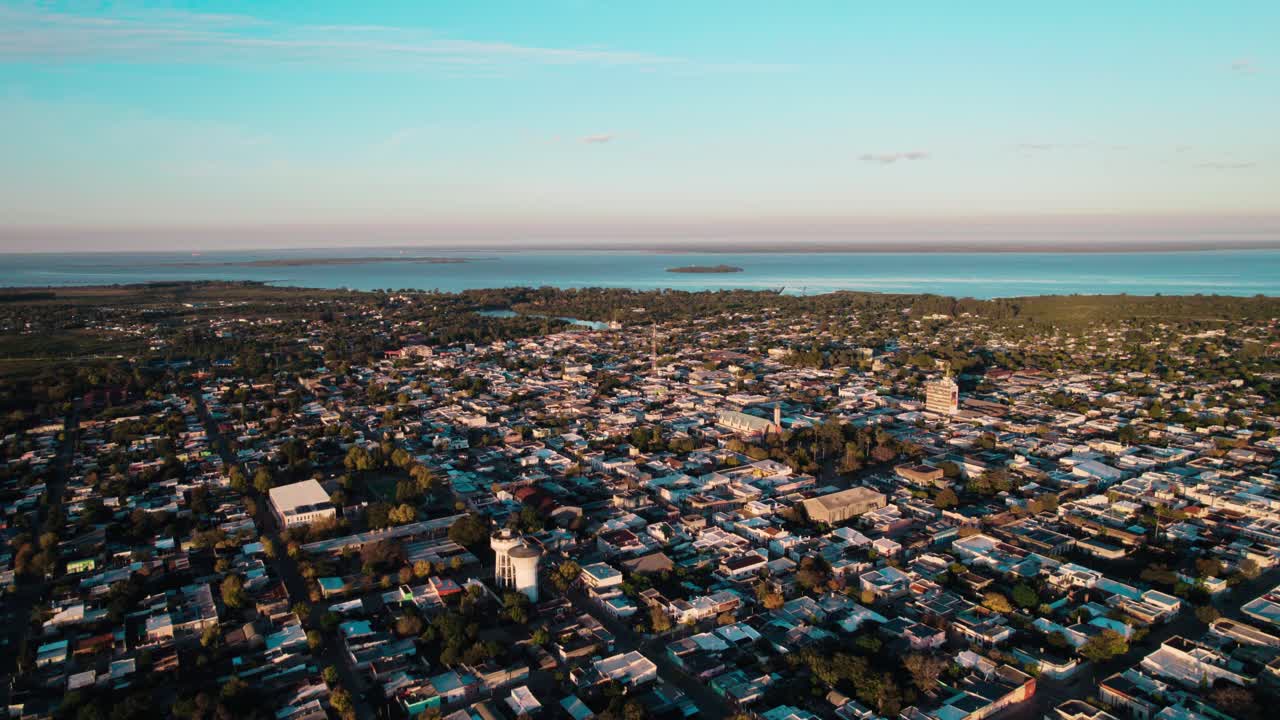 Drone shot capturing the dense residential and central area of Carmelo, Uruguay, extending toward the vast body of water (likely the Rio de la Plata)