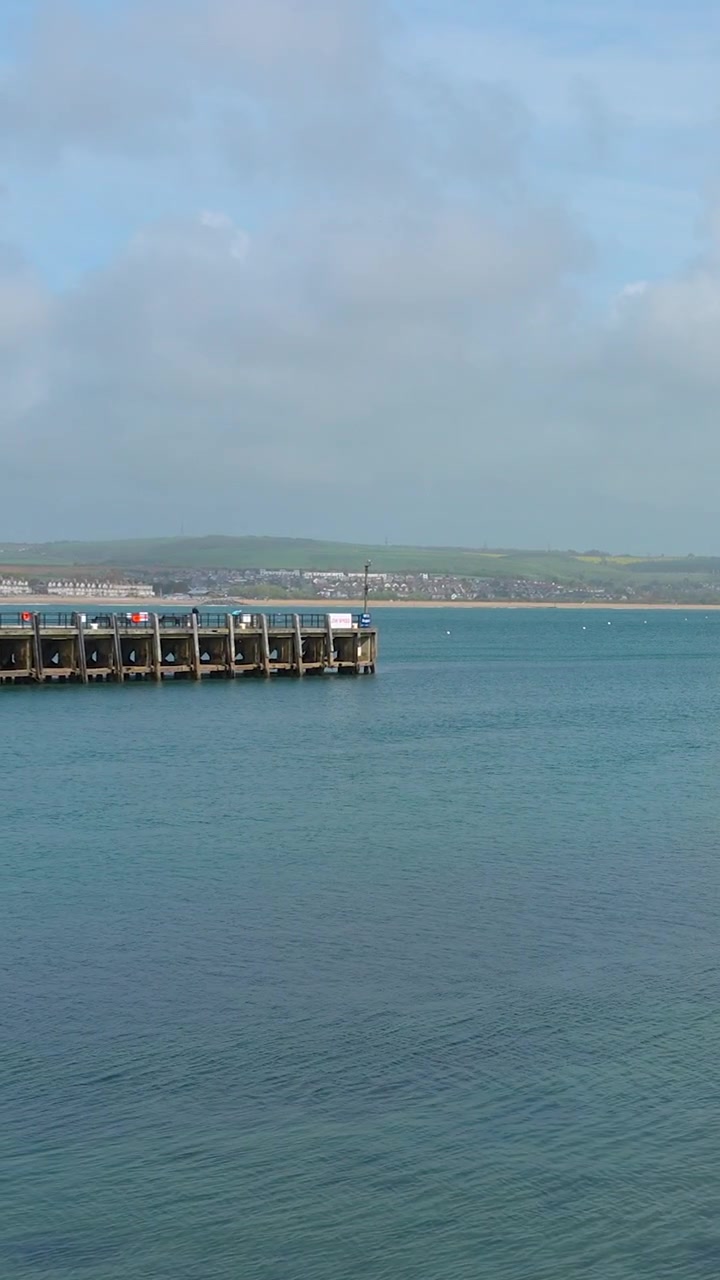 Uma tomada ampla do histórico cais do Porto de Weymouth, em Dorset, Inglaterra, contra um cenário de colinas verdes ondulantes e mar azul. Adequado para conteúdo de viagens e turismo