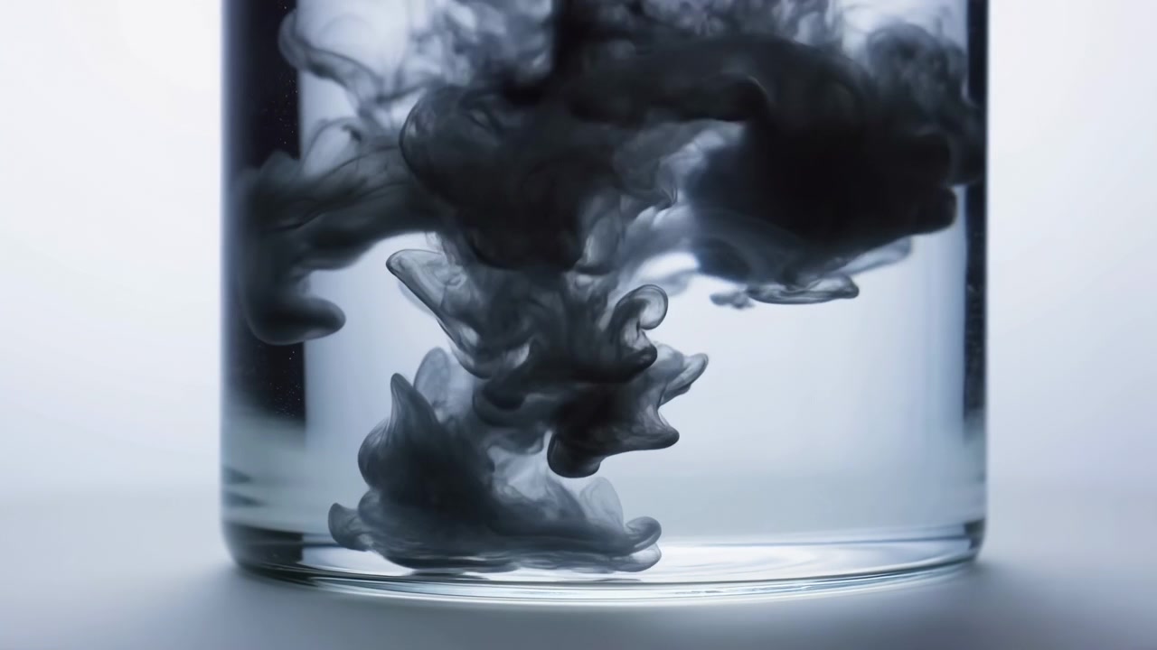 Dense charcoal ink droplet sinks and spreads through clear liquid inside cylindrical glass, forming layered cloud shapes with curling tendrils, soft folds, textured gradients, and stark monochrome