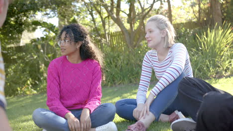 un grupo feliz y diverso de amigos adolescentes sentados en la hierba y hablando en un parque soleado, en cámara lenta