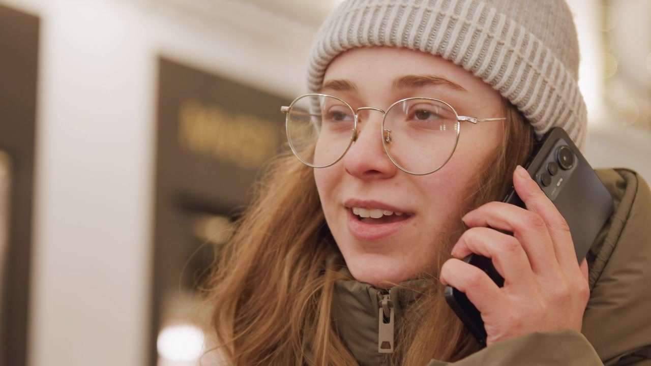 Closeup caucasian woman checking purse for phone then smiling during call in festive market setting, olive puffer jacket and beanie, eyeglasses, hands searching zipper, retrieves smartphone, dials