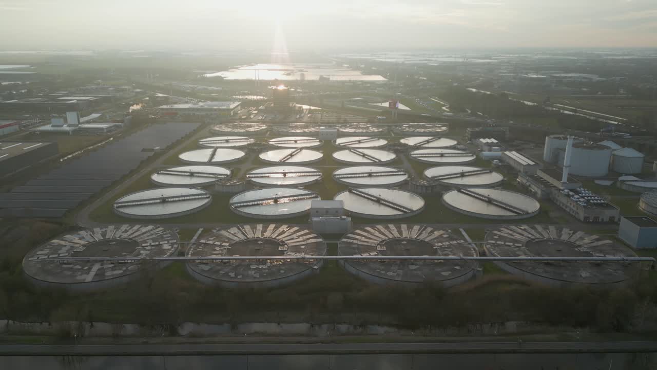 Wide drone shot showing solar panel rows in the foreground and circular wastewater treatment basins at the Harnaschpolder industrial water purification plant in the Netherlands