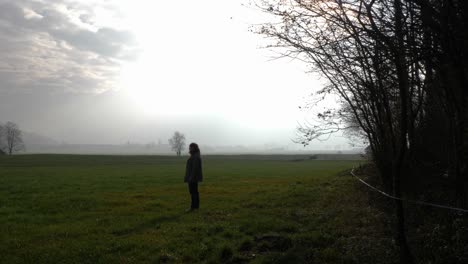 A-middle-aged-woman-with-a-brown-jacket-walks-on-a-green-gras-field-and-suddenly-stops-and-looks-around