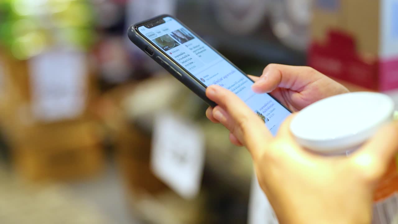 Hands holding a smartphone and coffee cup, scrolling through content in a vibrant market environment