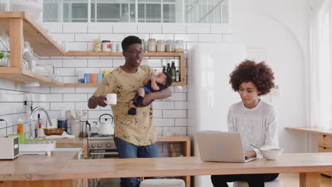 Busy-Family-In-Kitchen-At-Breakfast-With-Mother-Working-On-Laptop-And-Father-Caring-For-Baby-Son