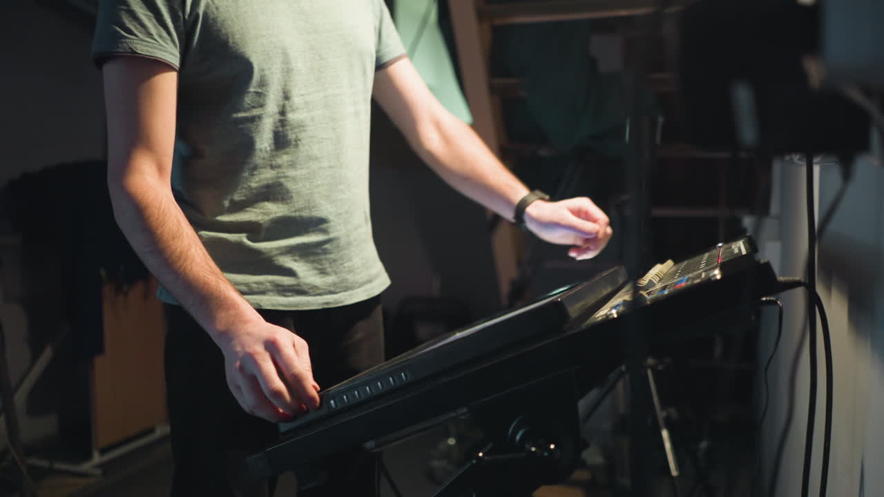 Young man in grey shirt stands under dim lighting in studio near metal staircase, looking down thoughtfully with serious expression, surrounded by industrial equipment and soft shadows creating moody atmosphere