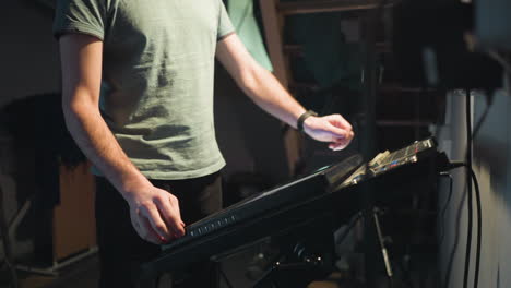 young man in grey shirt stands under dim lighting in studio near metal staircase, looking down thoughtfully with serious expression, surrounded by industrial equipment and soft shadows creating moody atmosphere