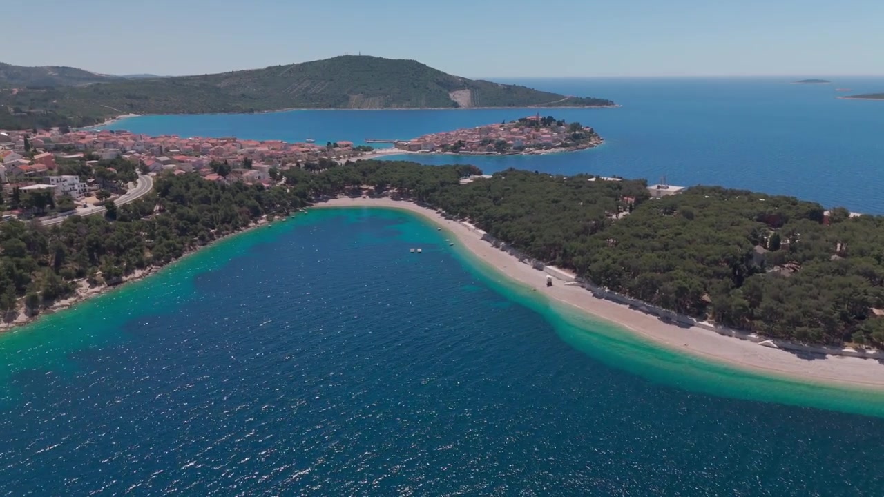 Dron se blíží k zalesněné pláži na poloostrově Primošten s širokými výhledy na Jadran, poté se plynule otáčí k městu a jeho pobřeží v jasném letním světle.