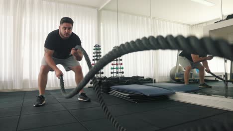 strong focused young bearded man using battle ropes for whipping exercise in a gym