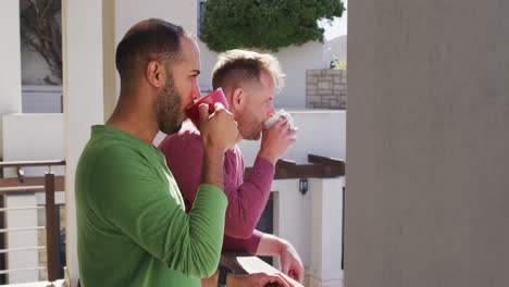 Multi-ethnic-gay-male-couple-drinking-coffee-on-balcony-in-sun