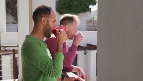 pareja de hombres homosexuales multiétnicos bebiendo café en el balcón al sol