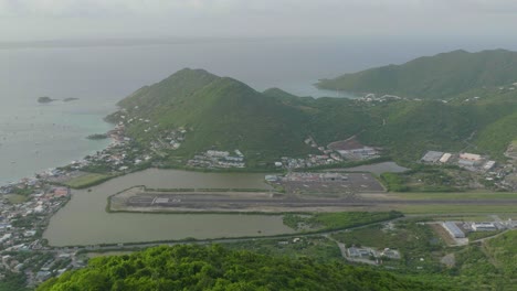 Aeria-moving-shot-of-Grand-Case-Airport-with-plane-departing-in-Saint-Martin-with-beautiful-Caribbean-landscape-at-background