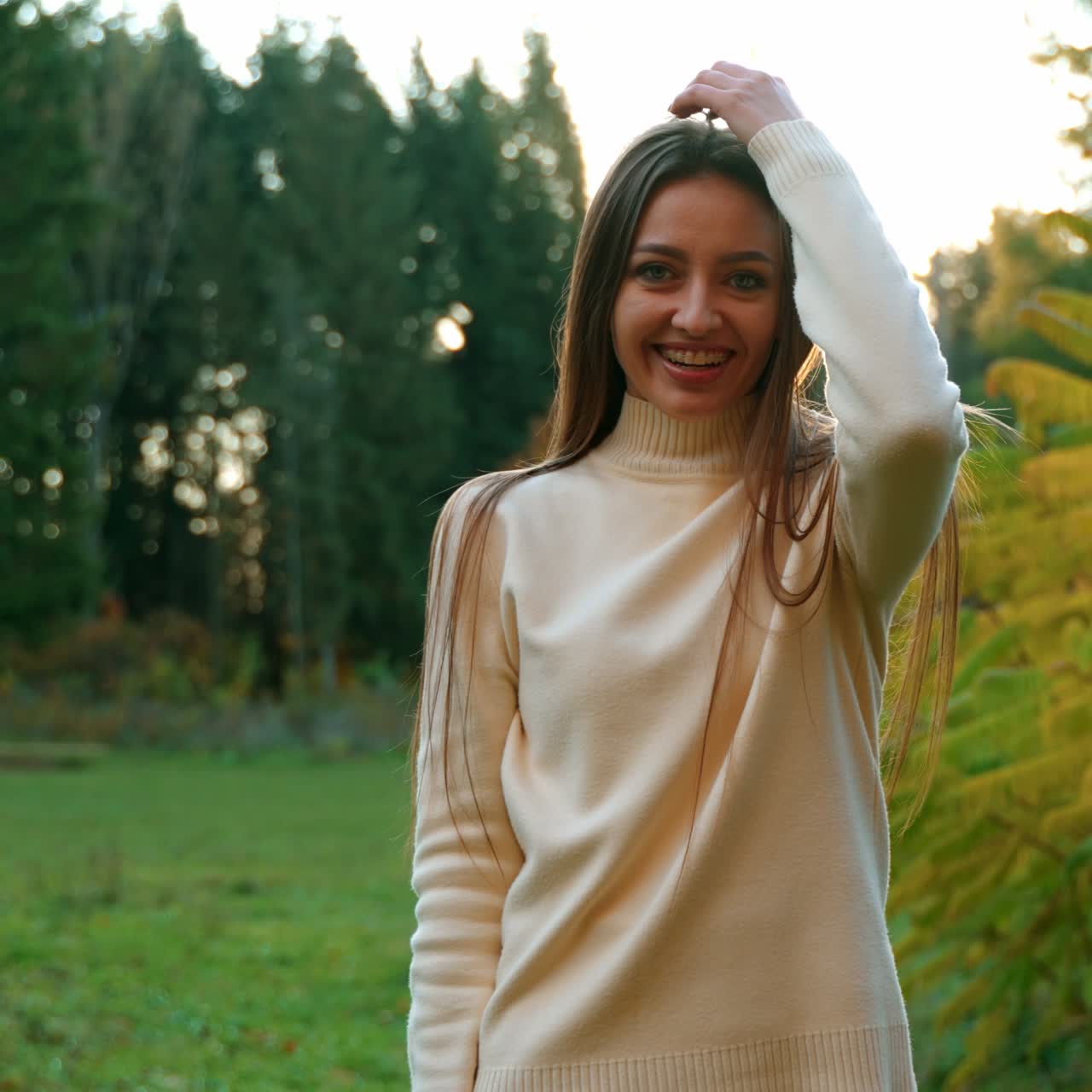 Happy smiling dark-haired woman waving her long hair in front of camera. Lady in white sweater posing at the backdrop of autumn park