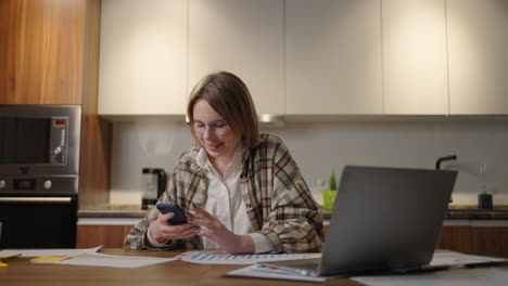 millennial woman employee sit by work desk chat in phone messenger app taking break in computer job. young lady worker manager browse web in mobile application on smartphone office from home