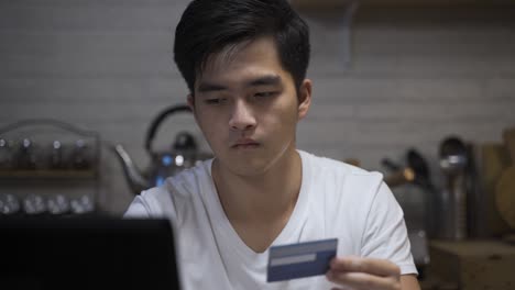 closeup shot of a cautious asian man is using a laptop to pay for taxes online with a credit card and a serious face in the dining room at home during nighttime