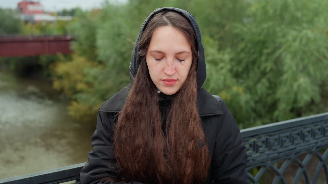Donna bianca in pausa meditativa su un ponte sul fiume, occhi chiusi, mani giunte, stato d'animo calmo e riflessivo, cielo grigio, sfondo verdeggiante, cappotto e cappuccio avvolgenti, pausa durante una passeggiata lenta, pensiero interiore e gentile solitudine