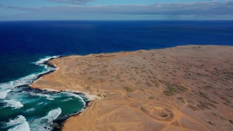 a drone surveys an arid headland riddled with off-road trails, where cobalt ocean swells hammer black rocks and white foam ribbons along the edge, conveying solitude and restless wind