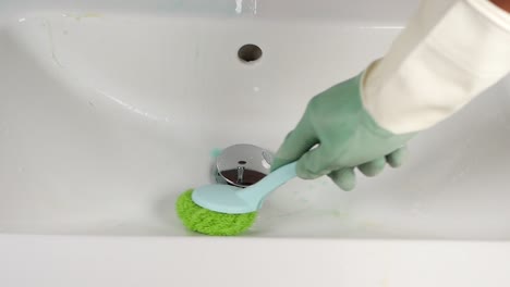 a hand wearing green gloves scrubs a white bathroom sink with a green and blue brush.