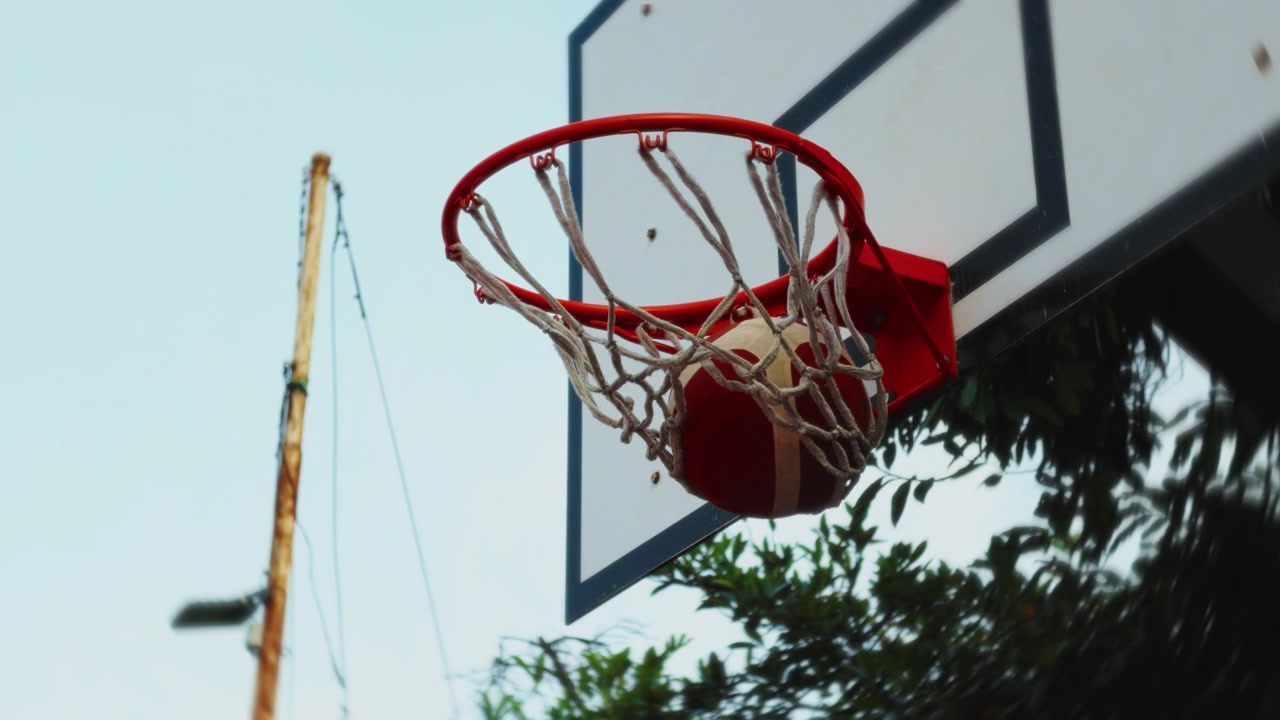 Pallone da basket che cade attraverso un canestro esterno al rallentatore, incorniciato da alberi e cielo.