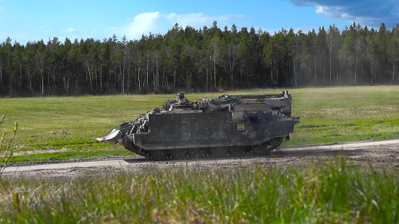British army or military Bulldog personnel carrier or tank vehicle that is heavily armored and has a bulldozer in front of it driving from grassy field to a gravel road creating dust in slow motion.