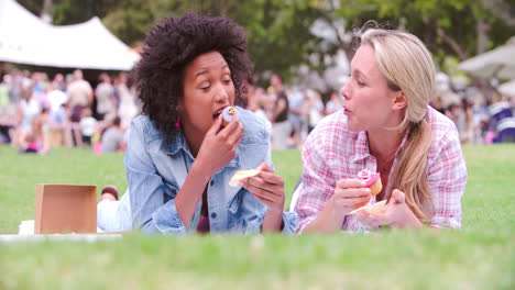 two friends lying on the grass and eating cakes at an event