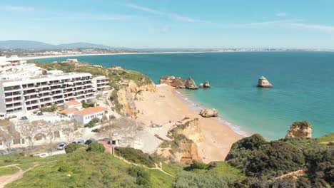 View-over-green-hill-shrouding-golden-Dona-Ana-beach,-Lagos,-Algarve,-Portugal---Aerial-Fly-over-Reveal-shot