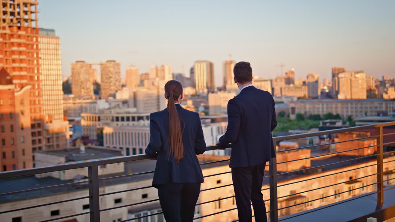 Office colleagues looking cityscape view on sunset rooftop. Two coworkers