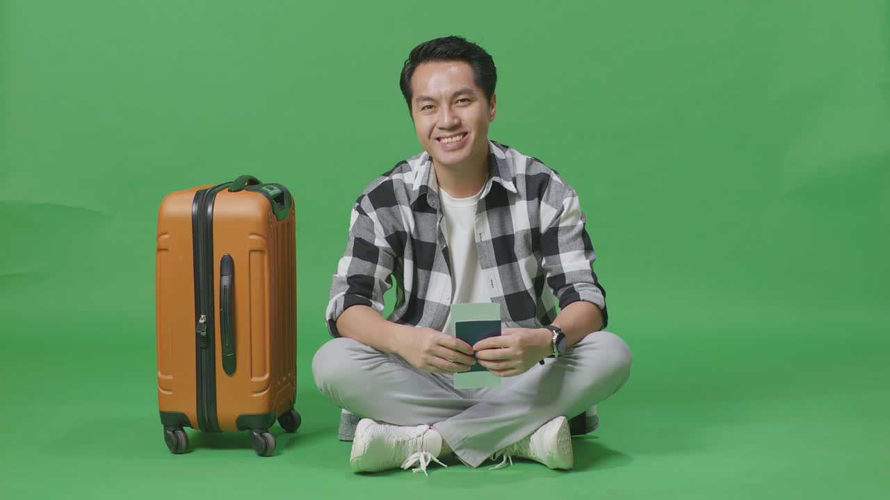 아시아 남성 여행자의 몸이 가방을 들고 여권을 들고 녹색 스크린 배경 스튜디오에 앉아있는 동안 카메라에 미소 짓고 있습니다.