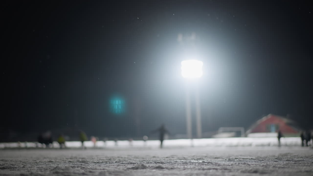 Blurred view of people skating and enjoying themselves on snow-covered outdoor ice rink illuminated by intense stadium lights during nighttime