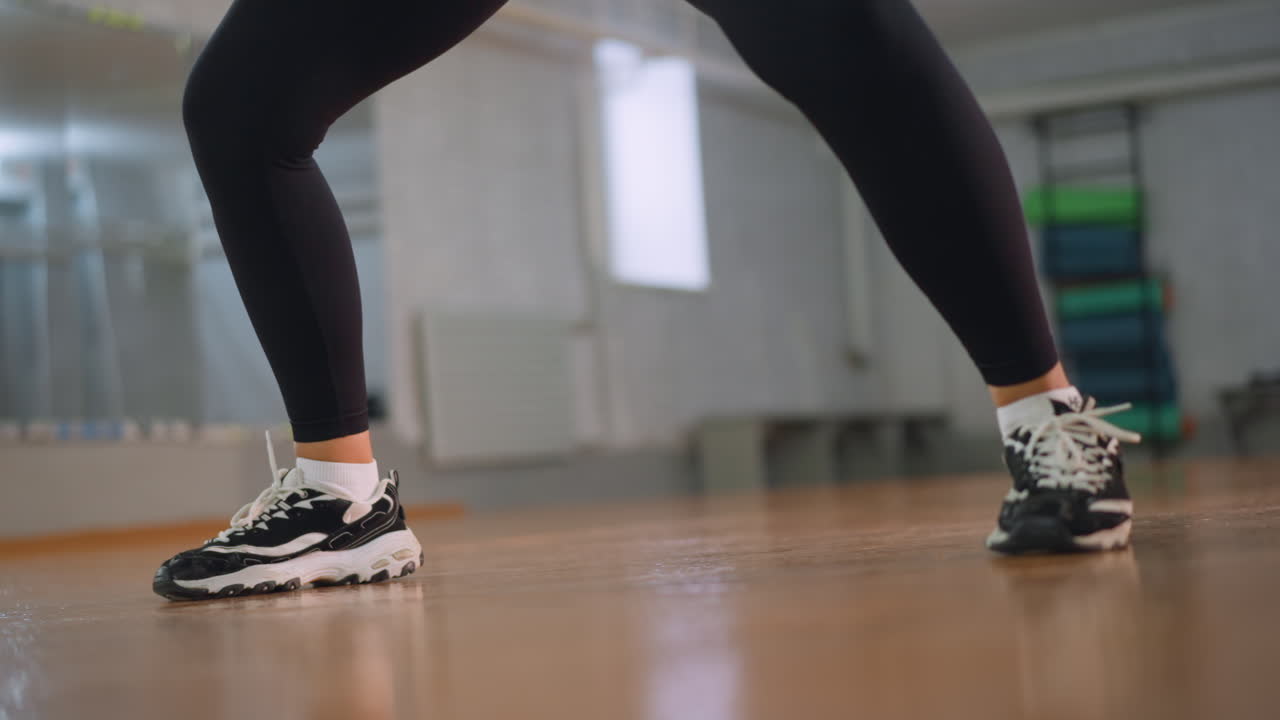 Leg view of woman in black leggings and sneakers bouncing on polished studio floor showing rhythm energy and motion during dance practice workout routine highlighting athletic movement strength