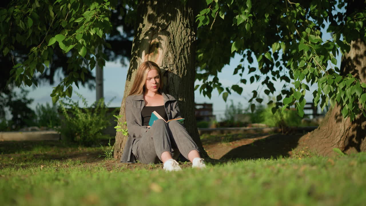 다리를 약간 펴고 야외에 앉아있는 여성, 잔디에 있는 나무에 기대어, 책을 읽고, 나무 잎이 바람에 부드럽게 흔들리고, 배경은 멀리 걸어가는 흐릿한 인물들을 보여줍니다.