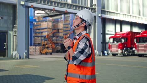 side view of asian male engineer with safety helmet pray for something while standing , outside of logistics distributions warehouse