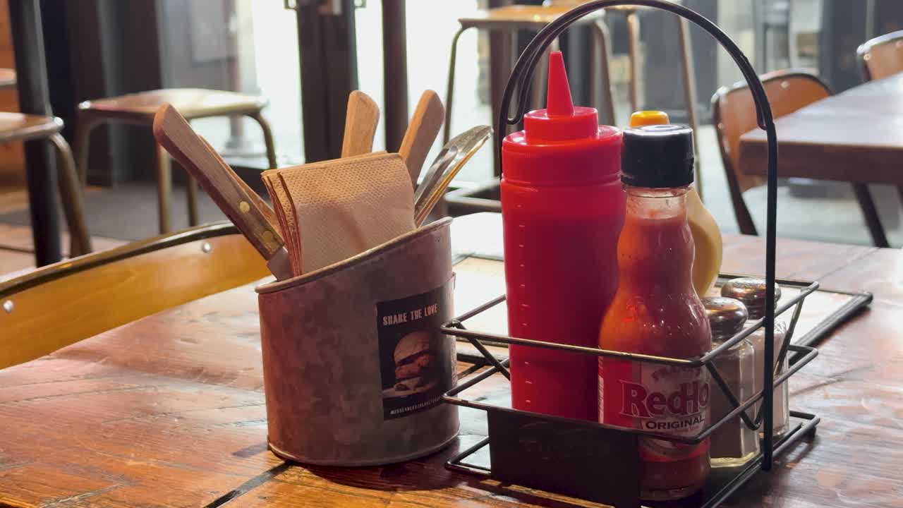 A hand adjusts a condiment holder on a wooden cafe table. Natural lighting highlights ketchup and sauce bottles, creating a casual dining atmosphere