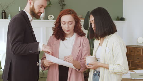 young redhead woman showing paper report to her two multiethnic colleagues and debating together 2