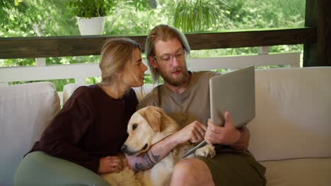 una chica rubia y un chico rubio con barba en gafas están mirando algo en una computadora portátil, un perro está sentado a su lado, que la chica está acariciando. ocio en el sofá en el gazebo en la naturaleza