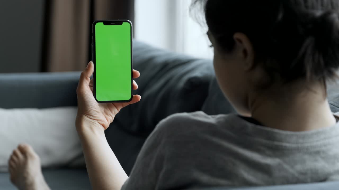 집에서 소파에 누워있는 여성은 녹색 모의 화면으로 스마트폰을 사용합니다. 소녀는 휴대 전화를 사용하고, 인터넷 소셜 네트워크를 탐색합니다. 시점 카메라.