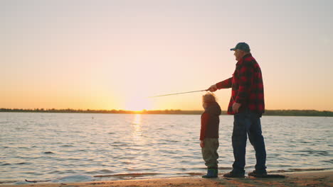 happy father and son are fishing on river shore in sunset old man and little child